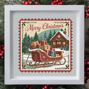 May include: Framed cross-stitch artwork depicting a Christmas scene. A red sleigh laden with presents is in the foreground, with a snow-covered cabin and fir trees in the background. The words "Merry Christmas" are at the top.