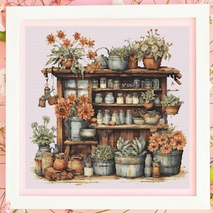 Puede incluir: Imagen de punto de cruz de un cobertizo rústico lleno de plantas en macetas, flores y tarros. La paleta de colores incluye tonos de marrón, naranja y verde, creando una escena cálida. La obra está enmarcada.
