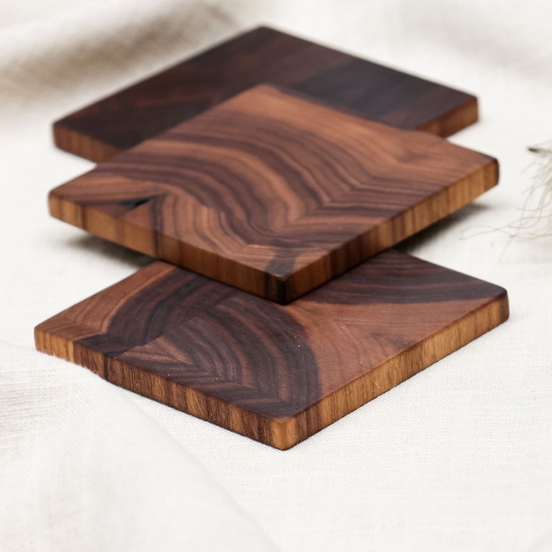 Drei quadratische Untersetzter aus Nussbaum Holz, arrangiert auf einem weißen Leinentuch. Die verschiedenen Brauntöne des Holzes sowie die glatte Oberfläche sind ersichtlich.