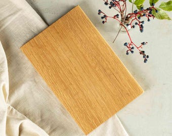 May include: A rectangular wooden chopping board with a light brown finish. The board is positioned diagonally on a beige linen cloth. A small glass vase with berries and greenery is in the background.