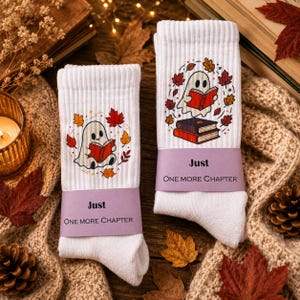 Könnte beinhalten: Ein Paar weiße gerippte Socken mit einer Grafik eines Geistes, der ein rotes Buch liest, umgeben von Herbstblättern. Ein violettes Papierband umschließt die Mitte jeder Socke mit dem aufgedruckten Text Just ONE MORE CHAPTER.