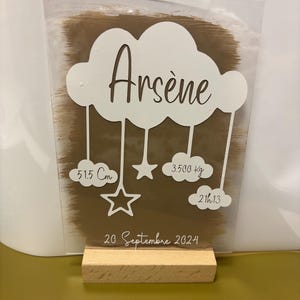 Puede incluir: Una placa acrílica blanca con un fondo marrón y una nube blanca con el nombre "Arsène" escrito en marrón. La nube está colgada de tres cuerdas con estrellas y nubes adjuntas. La placa también incluye la fecha "20 Septembre 2024", la hora "21h13", el peso "3.500 kg" y la longitud "51.5 cm".