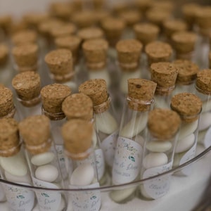 Puede incluir: Un tarro de vidrio transparente lleno de pequeños frascos de vidrio, cada uno con un tapón de corcho y lleno de caramelos blancos y verdes. Los frascos tienen etiquetas con el texto "Elise & Drew" y la fecha "8 Juin 2024".