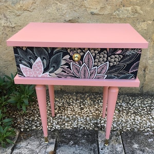 Könnte beinhalten: Ein rosa Nachttisch mit einer einzigen Schublade mit einem schwarzen und rosa Blumendruck. Die Schublade hat einen goldenen Knopf. Der Tisch hat vier konische Beine.
