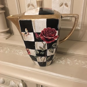 Könnte beinhalten: Eine dekorative Teekanne mit einem schwarz-weißen Schachbrettmuster, goldenen Verzierungen und einem roten Rosendesign. Die Teekanne hat einen Deckel und einen Griff. Auf der Seite ist auch ein Schmetterling gemalt.