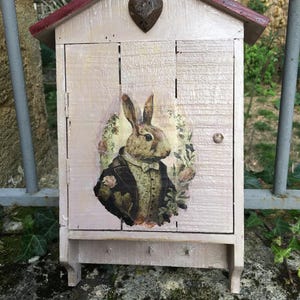 May include: A light pink wooden key cabinet with a red roof. It features a vintage illustration of a rabbit in a suit. A metal heart decorates the top. The cabinet has small hooks for keys.