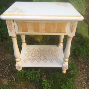 Op de afbeelding: Witte bijzettafel met gouden accenten en een decoratief patroon op de bovenste en onderste planken. De tafel heeft een lade met een goud-wit gestreept ontwerp.