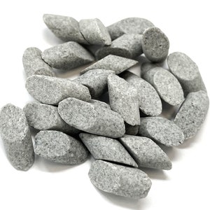May include: A pile of small, gray, oblong stones with a speckled texture. The stones are roughly the same size and shape, with rounded ends and a flattened, diamond-like center. The stones are scattered on a white surface.