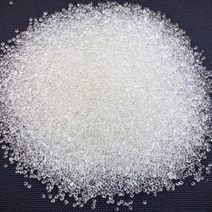 May include: A close-up shot of a pile of small, clear, spherical beads. The beads are densely packed together, creating a textured surface. The background is a dark, solid color, providing contrast to the translucent beads.