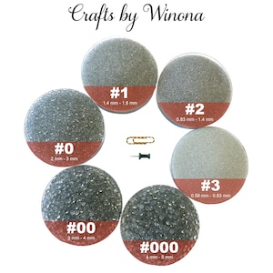 Op de afbeelding: Zes doorzichtige glazen potten gevuld met verschillende maten glazen kralen. De potten zijn gelabeld met de nummers 0, 00, 1, 2, 3 en 000. Elke pot heeft een label met de overeenkomstige maat van de kralen in millimeters.