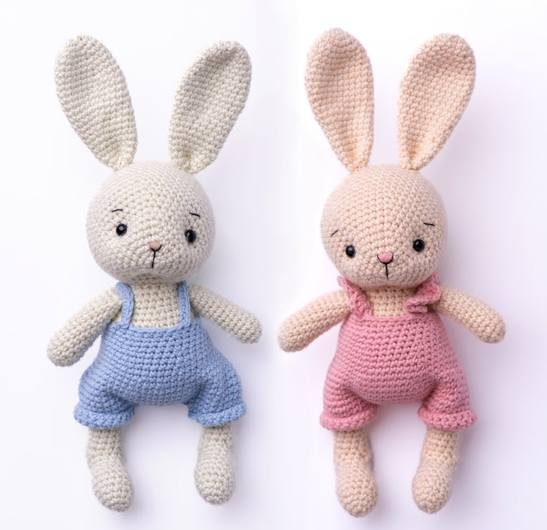 Op de afbeelding: Twee gehaakte konijnen speelgoed, een met een blauwe overall en een met een roze overall. Beide konijnen hebben beige vacht en zwarte ogen.