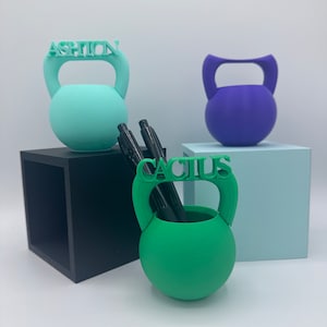 以下が含まれることがあります： 緑、青、紫のケトルベル型のデスクオーガナイザー3つ。緑には「FASHION」、紫には「CACTUS」と書いてあり、紫には何も書いてありません。緑には黒いペンが2本入っています。