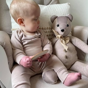 May include: A baby wearing a light brown onesie and pants sits in a rocking chair next to a stuffed teddy bear. The teddy bear has a light brown body, dark brown ears, and a tan bow. The baby is holding a wooden toy.