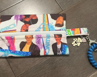 Könnte beinhalten: Eine rechteckige Reißverschlusstasche aus Stoff mit einem wiederkehrenden Druck der Mitglieder von New Kids on the Block samt Namen, einem weißen Reißverschluss, einem kleinen silbernen Roboteranhänger und einem blauen Spiralarmband.