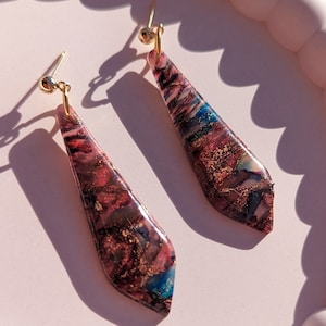 May include: A pair of dangle earrings with gold hooks and teardrop-shaped resin pendants. The resin is translucent and features a swirling pattern of pink, blue, and gold.