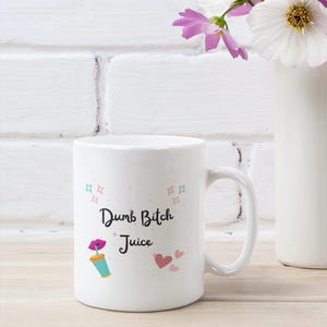 May include: White ceramic mug with the text "Dumb Bitch Juice" in black script, accompanied by a cartoon image of a lip, a drink, and hearts. The mug has a curved handle and is set against a white brick wall.