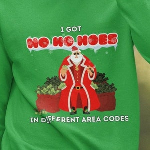 May include: Green sweatshirt with the text "I GOT HO HO HOES IN DIFFERENT AREA CODES." The graphic features Santa Claus in a red coat, sunglasses, and holding money, with bags of money on either side.