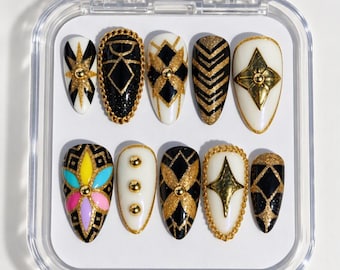 Pode incluir: Um conjunto de dez unhas postiças em formato de amêndoa exibidas num estojo de plástico transparente. Os designs apresentam uma mistura de padrões pretos, brancos e glitter dourado, incluindo formas geométricas, tachas douradas, bordas de corrente e flores.
