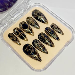 Puede incluir: Un juego de diez uñas postizas negras y doradas. Cada uña presenta un diseño dorado único, que incluye patrones geométricos y motivos de sol. Las uñas tienen forma de almendra y se exhiben en una caja de plástico transparente con interior beige.