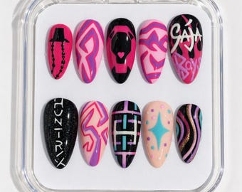 May include: A set of ten almond-shaped press-on nails in a clear plastic case. The designs feature black, pink, purple, and peach colors with patterns including graffiti-style text reading "GAJA BOYS" and "HUNTR/X", geometric lines, a star, and dots.