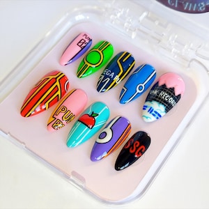 May include: A set of ten long, oval-shaped artificial nails with various designs. The nails feature bright colors like pink, green, blue, and red, with patterns including stripes, circles, and text. The nails are displayed in a clear plastic case.