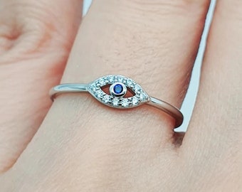 May include: A silver ring featuring an eye design. The eye is outlined with small, clear stones and has a blue center. The ring is on a finger, showcasing the delicate details of the jewelry.