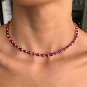 Könnte beinhalten: Eine goldene Halskette mit einer Reihe von tropfenförmigen, tiefroten Edelsteinen. Die Halskette wird um den Hals getragen und zeigt die leuchtende Farbe und das aufwendige Design des Schmucks.