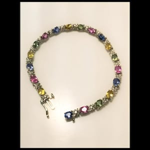 Puede incluir: Una pulsera de plata con una serie de piedras preciosas multicolores en tonos azules, rosas, amarillos y verdes. Las piedras de talla ovalada están engastadas en una banda de plata con un cierre. La pulsera se muestra sobre una superficie blanca.