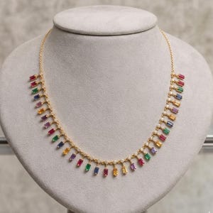 May include: A gold necklace with a series of colorful rectangular gemstones. The gemstones are red, green, blue, yellow, and purple. The necklace is displayed on a gray bust.