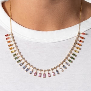 May include: A gold chain necklace with a rainbow of baguette-shaped sapphires hanging from it. The sapphires are arranged in a graduated pattern, with the largest sapphires at the bottom of the necklace. The necklace is a beautiful and unique piece of jewelry.