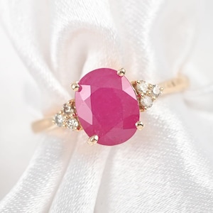 May include: A gold ring featuring a large, oval-cut ruby as the centerpiece. The ruby is a vibrant pink-red color. Small diamonds are clustered on either side of the ruby, adding sparkle. The ring is set against a white fabric background.