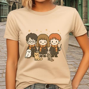 Può includere: T-shirt beige con un'illustrazione a cartoni animati di tre personaggi con occhiali, sciarpe e vesti, insieme a un gufo, un gatto e un topo. Lo sfondo mostra un edificio in pietra. La maglietta è di colore marrone chiaro.