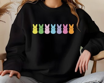 Peut inclure: Sweat-shirt noir avec une rangée de guimauves en forme de lapin de couleurs vives. Les lapins sont dans un ordre de couleurs arc-en-ciel, commençant par le jaune et se terminant par l'orange.