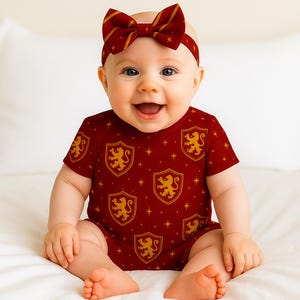 Puede incluir: Un bebé con un body burdeos y una diadema a juego, ambos con un estampado de escudos de león dorados. El body tiene mangas cortas. La diadema tiene un lazo. El bebé está sonriendo.