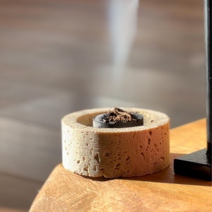 Puede incluir: Primer plano de un quemador de incienso redondo de color beige texturizado con un disco de carbón negro en su interior. El humo se eleva del disco de carbón.
