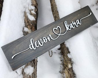 May include: A rectangular wooden sign with the names "devon" and "kara" in white cursive script, connected by a heart symbol. The sign is a dark gray color and rests on a snowy surface.