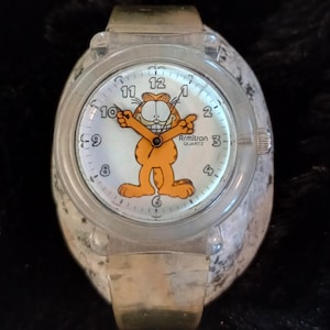 May include: A clear plastic watch with a gold metal band. The watch face features a cartoon image of Garfield the cat with the text "Armitron Quartz".