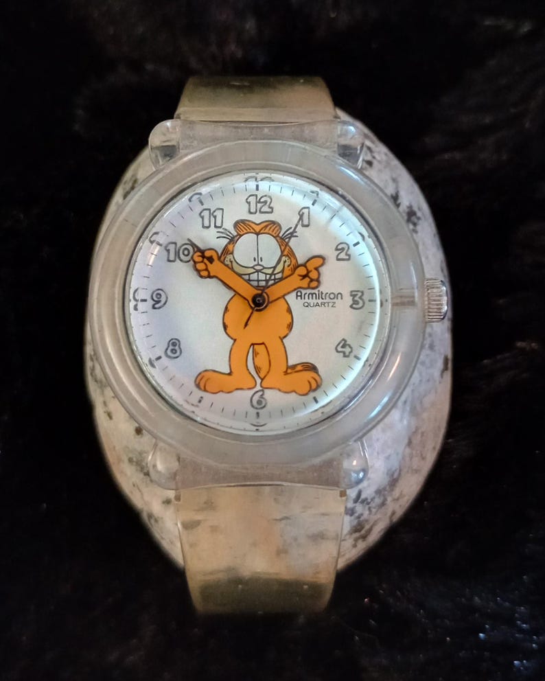 May include: A clear plastic watch with a gold metal band. The watch face features a cartoon image of Garfield the cat with the text "Armitron Quartz".
