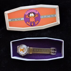May include: A clear plastic watch with a Garfield cartoon character on the face. The watch is in a white box with a purple interior. The box has the words "Garfield Collectibles" and "Armitron" on it.