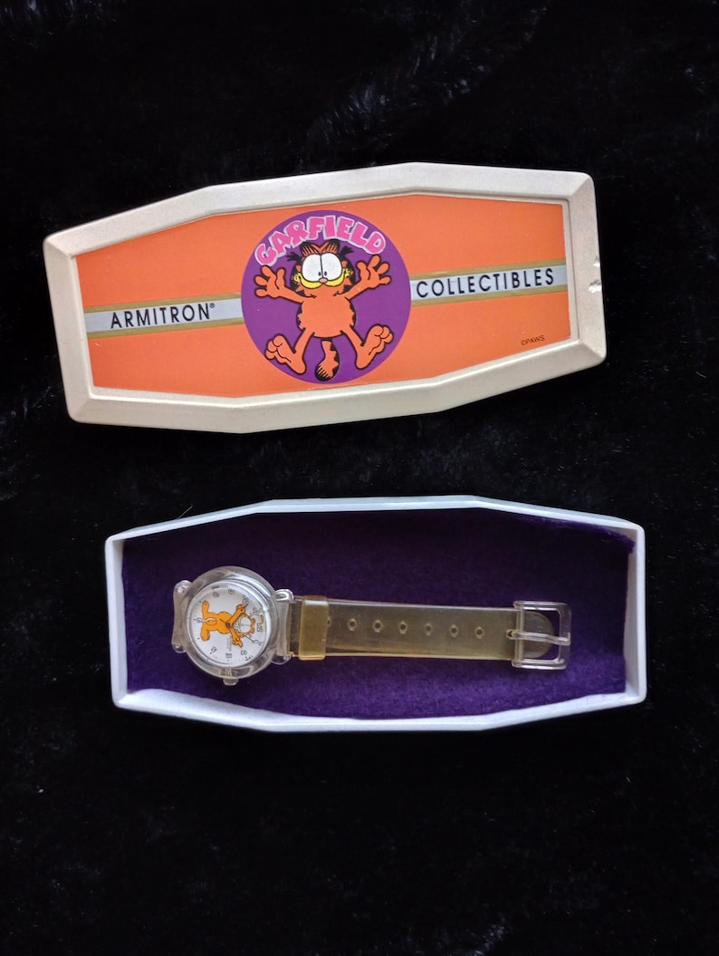 May include: A clear plastic watch with a Garfield cartoon character on the face. The watch is in a white box with a purple interior. The box has the words "Garfield Collectibles" and "Armitron" on it.