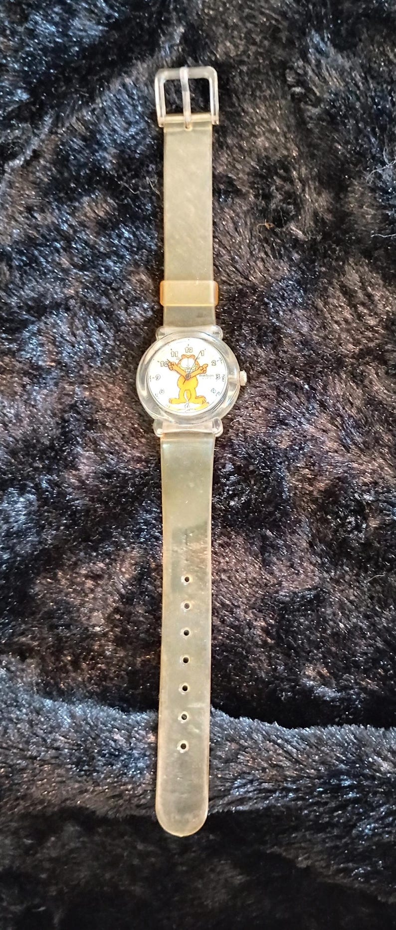 May include: A clear plastic watch with a white face featuring a cartoon image of Garfield. The watch has a clear plastic band with a silver buckle.
