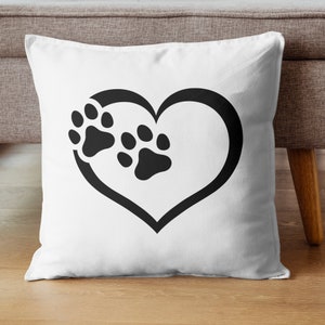May include: White square throw pillow featuring a black heart outline with two black paw prints inside. The pillow is on a wooden floor, and the background includes a gray sofa and a wooden table leg.