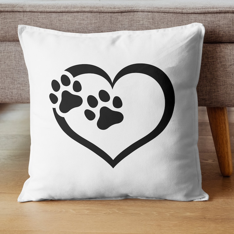 May include: White square throw pillow featuring a black heart outline with two black paw prints inside. The pillow is on a wooden floor, and the background includes a gray sofa and a wooden table leg.
