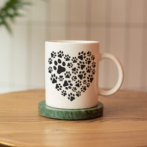 May include: White ceramic mug featuring a heart design made of black paw prints. The mug sits on a green marble coaster, placed on a wooden surface. The mug has a curved handle and is suitable for hot or cold beverages.