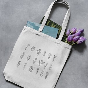 May include: A white canvas tote bag with black line drawings of flowers. The bag is filled with a magazine and a bouquet of purple tulips.