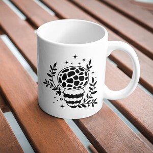 May include: White ceramic mug with a black and white illustration of a mushroom with a patterned cap and stem. The mushroom is surrounded by leaves and stars.