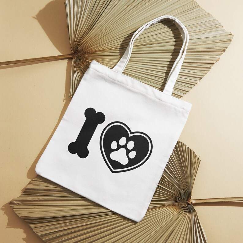 May include: White tote bag with black graphic of a bone, a heart, and a paw print. The bag has two white handles and is set against a neutral background with dried palm leaves.