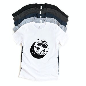 May include: White t-shirt with a black and white graphic of a mushroom growing from a crescent moon with stars. The shirt is layered on top of other t-shirts in various shades of gray and blue.