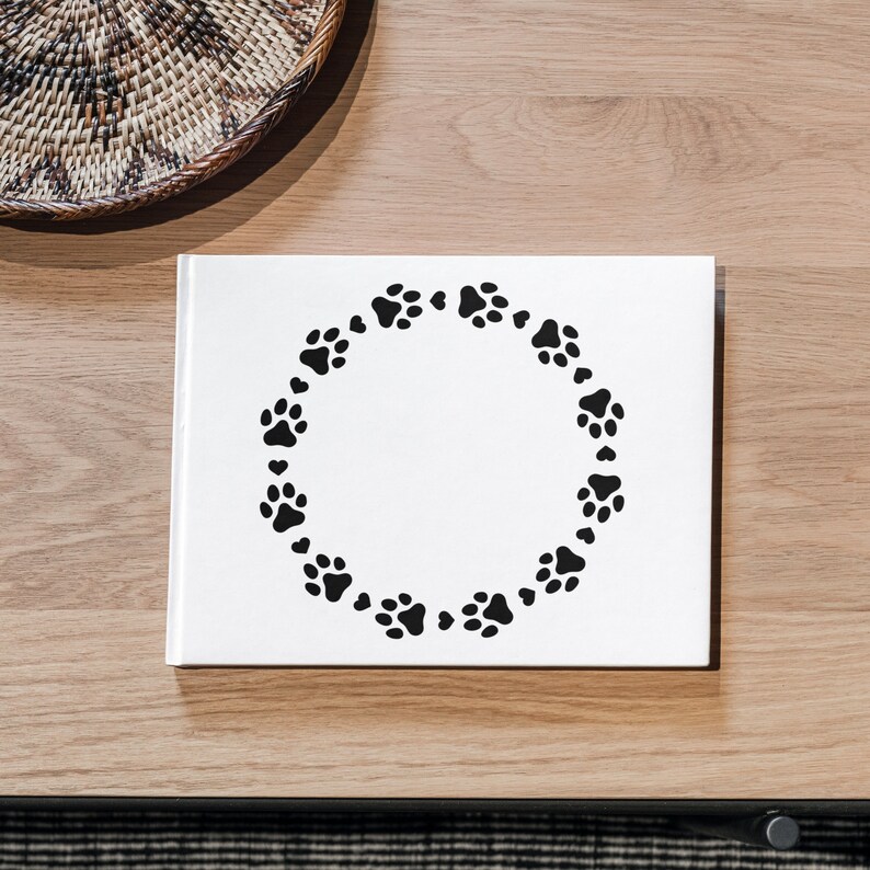 May include: A white book with a black paw print circle design. The paw prints are arranged in a circular pattern, creating a decorative border. The book is on a wooden surface, with a woven basket in the background.
