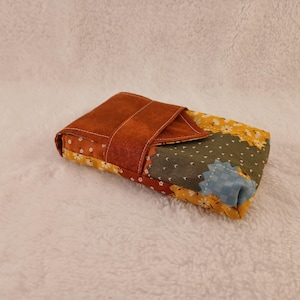 May include: A rectangular, patchwork fabric case with a brown leather flap closure. The case features a mix of floral and solid fabrics in shades of yellow, green, orange, and blue. The case is designed to hold small items.